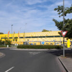 Außenansicht eines gelben DHL-Depots der Deutschen Post mit Lieferfahrzeugen und Zufahrtsstraße.