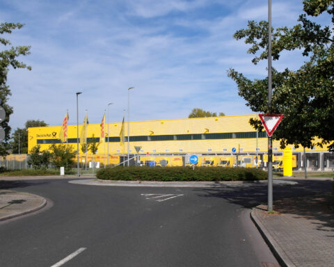 Außenansicht eines gelben DHL-Depots der Deutschen Post mit Lieferfahrzeugen und Zufahrtsstraße.