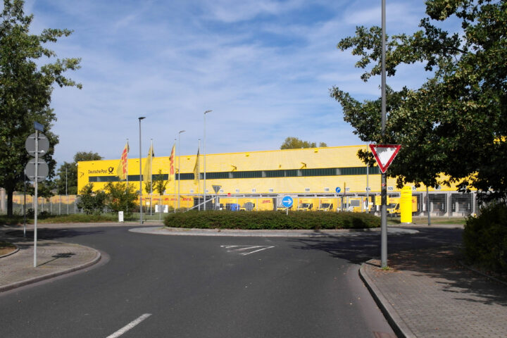 Außenansicht eines gelben DHL-Depots der Deutschen Post mit Lieferfahrzeugen und Zufahrtsstraße.