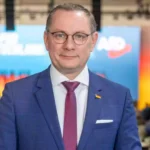 Tino Chrupalla steht in einem Veranstaltungsraum vor einem AfD-Hintergrund.