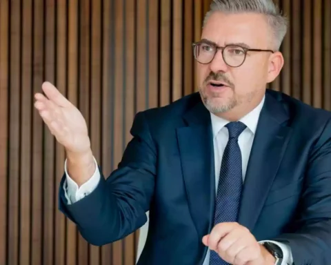 Ein Mann im dunklen Anzug mit Brille gestikuliert bei einem Gespräch vor einer Holzlamellenwand.