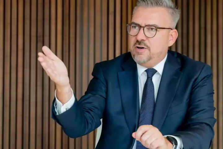 Ein Mann im dunklen Anzug mit Brille gestikuliert bei einem Gespräch vor einer Holzlamellenwand.
