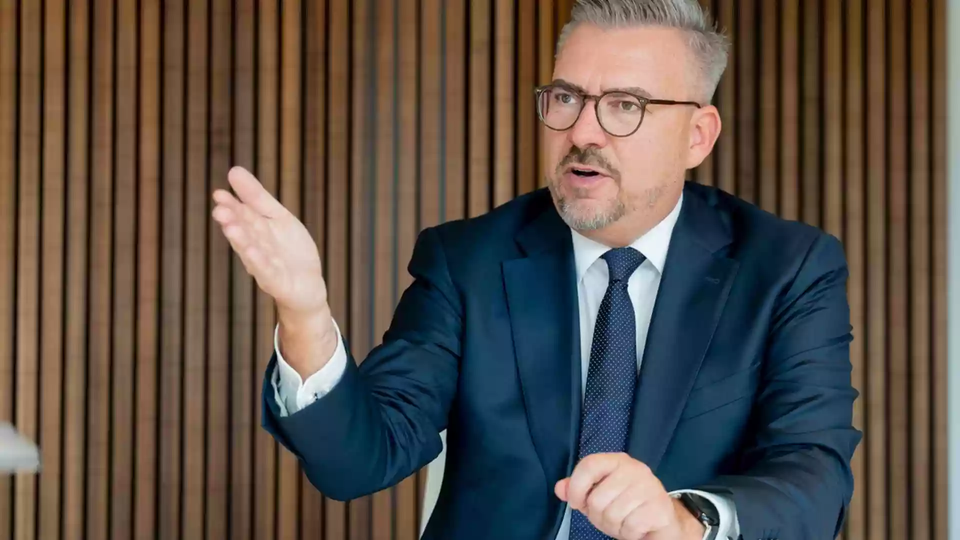 Ein Mann im dunklen Anzug mit Brille gestikuliert bei einem Gespräch vor einer Holzlamellenwand.