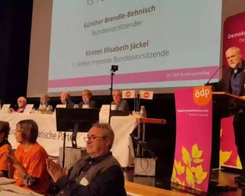 Redner am Podium auf einem ÖDP-Parteitag vor einer Bühne mit Parteibannern und Delegierten.