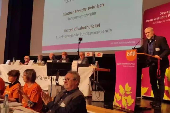 Redner am Podium auf einem ÖDP-Parteitag vor einer Bühne mit Parteibannern und Delegierten.