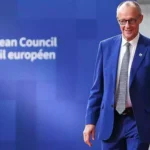 Friedrich Merz steht vor einer blauen European-Council-Wand neben einer EU-Flagge.