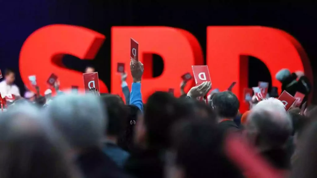 Menschen halten auf einem SPD-Parteitag rote Abstimmungskarten vor großen roten SPD-Buchstaben.