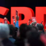 Menschen halten auf einem SPD-Parteitag rote Abstimmungskarten vor großen roten SPD-Buchstaben.