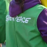 Person mit grünem Greenpeace-Überwurf und violettem Kapuzenpullover von hinten.