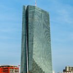 Modernes Glas-Hochhaus an einem klaren Himmel, davor Uferpromenade mit Bäumen und einzelnen Gebäuden.