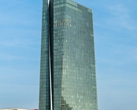 Modernes Glas-Hochhaus an einem klaren Himmel, davor Uferpromenade mit Bäumen und einzelnen Gebäuden.