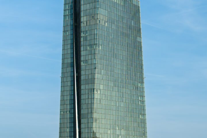 Modernes Glas-Hochhaus an einem klaren Himmel, davor Uferpromenade mit Bäumen und einzelnen Gebäuden.