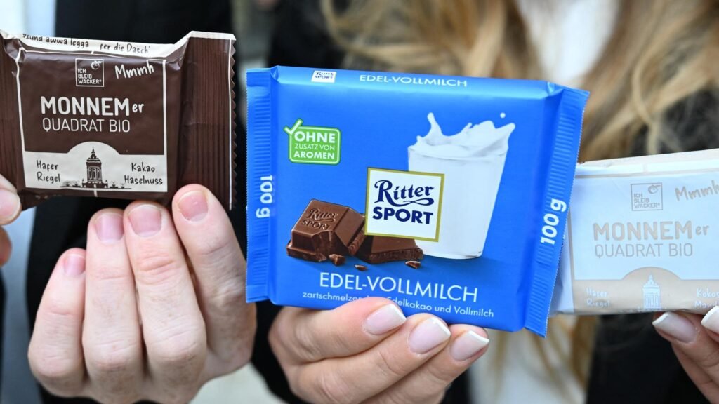 Hände halten zwei Ritter-Sport-Schokoladentafeln und eine Wacker-Verpackung mit der Aufschrift „Monnemer Quadrat Bio“.