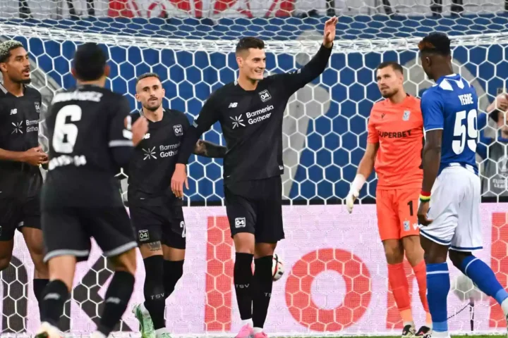 Bayer-Leverkusen-Spieler feiern vor dem Tor, während der gegnerische Torhüter und ein Verteidiger zuschauen.