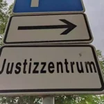 Straßenschild mit der Aufschrift „Justizzentrum“ und Pfeil nach rechts vor Bäumen.