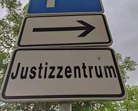 Straßenschild mit der Aufschrift „Justizzentrum“ und Pfeil nach rechts vor Bäumen.