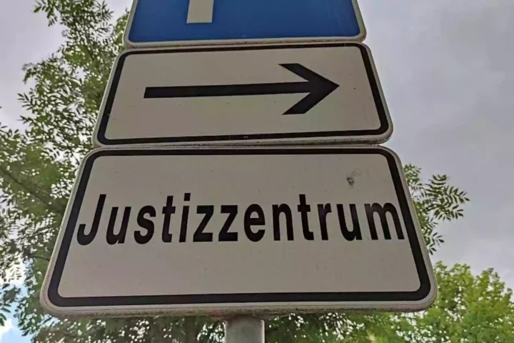 Straßenschild mit der Aufschrift „Justizzentrum“ und Pfeil nach rechts vor Bäumen.