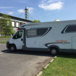 Ein Wohnmobil der Marke Weinsberg steht vor einem Stadiongebäude unter blauem Himmel.