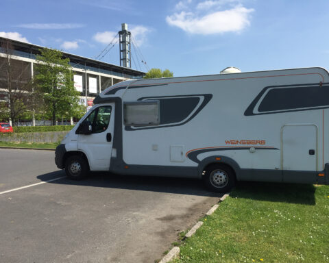 Ein Wohnmobil der Marke Weinsberg steht vor einem Stadiongebäude unter blauem Himmel.