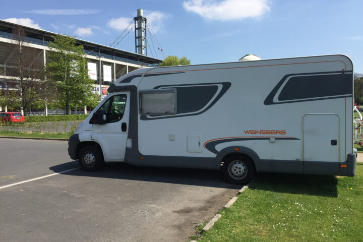 Ein Wohnmobil der Marke Weinsberg steht vor einem Stadiongebäude unter blauem Himmel.