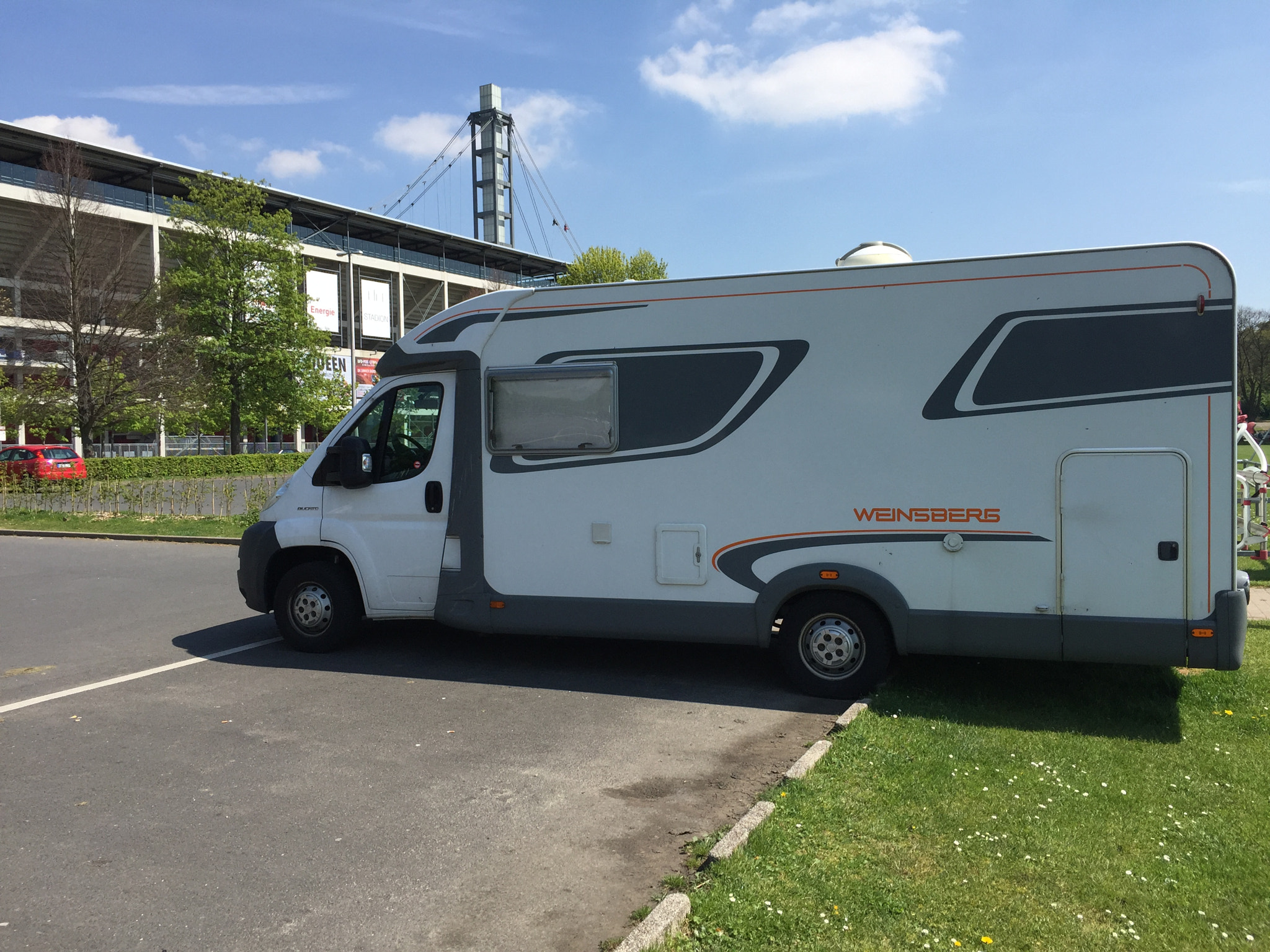 Ein Wohnmobil der Marke Weinsberg steht vor einem Stadiongebäude unter blauem Himmel.