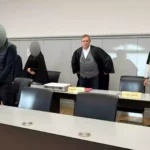 Mehrere Angeklagte und ein Richter stehen in einem Gerichtssaal.