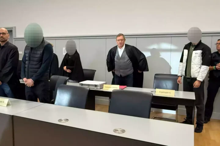 Mehrere Angeklagte und ein Richter stehen in einem Gerichtssaal.