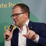 Ein Mann im dunkelblauen Anzug spricht bei einer Veranstaltung in ein Mikrofon und hebt den Zeigefinger.