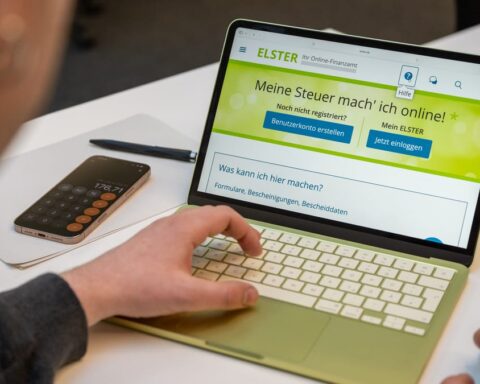 Person uses a laptop showing the ELSTER online tax portal next to a smartphone on a desk.