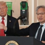 Donald Trump steht im Hintergrund, während ein Mann an einem Rednerpult vor Mikrofonen spricht und eine Geste mit ausgestrecktem Arm macht.