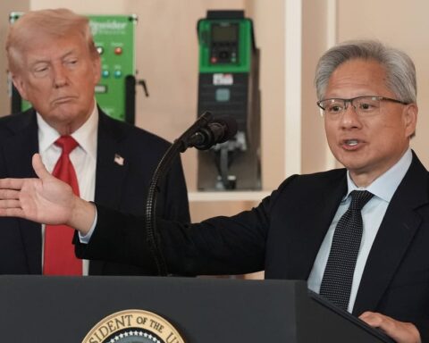 Donald Trump steht im Hintergrund, während ein Mann an einem Rednerpult vor Mikrofonen spricht und eine Geste mit ausgestrecktem Arm macht.