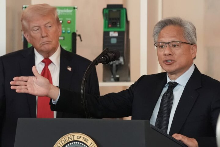 Donald Trump steht im Hintergrund, während ein Mann an einem Rednerpult vor Mikrofonen spricht und eine Geste mit ausgestrecktem Arm macht.