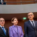 Drei Personen in Anzügen und einem lilafarbenen Blazer stehen in einem Sitzungssaal des Bundestags.