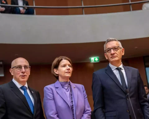 Drei Personen in Anzügen und einem lilafarbenen Blazer stehen in einem Sitzungssaal des Bundestags.