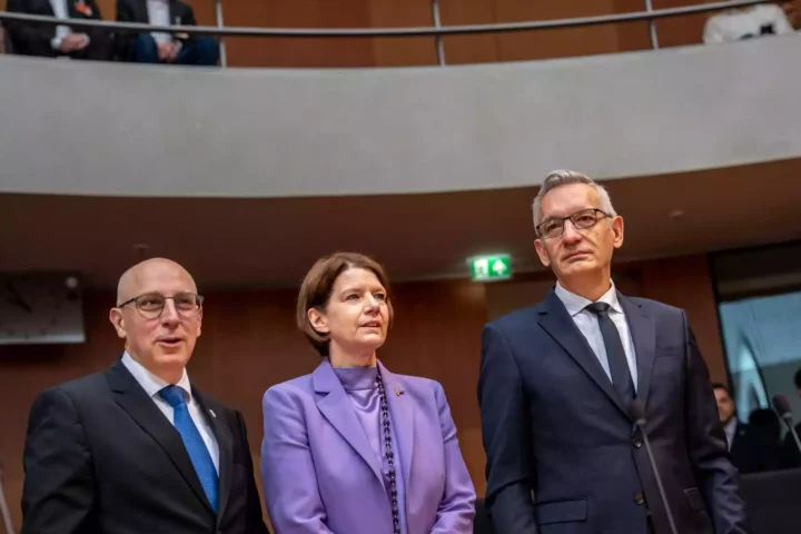 Drei Personen in Anzügen und einem lilafarbenen Blazer stehen in einem Sitzungssaal des Bundestags.