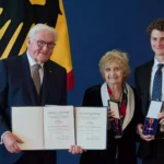Frank-Walter Steinmeier steht mit zwei Geehrten vor einer deutschen Flagge und hält Verleihungsurkunden zum Bundesverdienstkreuz.