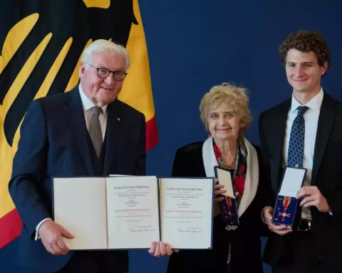 Frank-Walter Steinmeier steht mit zwei Geehrten vor einer deutschen Flagge und hält Verleihungsurkunden zum Bundesverdienstkreuz.