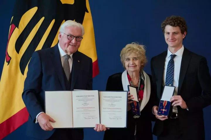 Frank-Walter Steinmeier steht mit zwei Geehrten vor einer deutschen Flagge und hält Verleihungsurkunden zum Bundesverdienstkreuz.