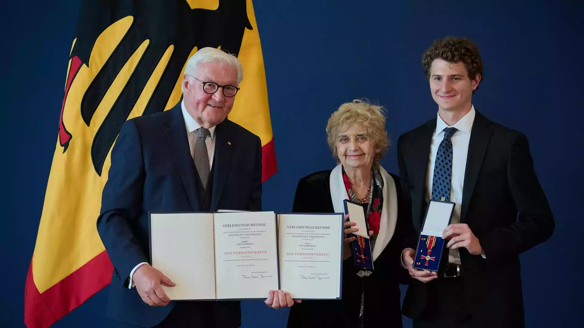 Steinmeier zeichnet 21 Menschen mit dem Bundesverdienstkreuz aus 1 Frank-Walter Steinmeier steht mit zwei Geehrten vor einer deutschen Flagge und hält Verleihungsurkunden zum Bundesverdienstkreuz.