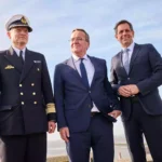 Drei Männer, darunter ein Marineoffizier und zwei Politiker in Anzügen, stehen am Wasser vor blauem Himmel.