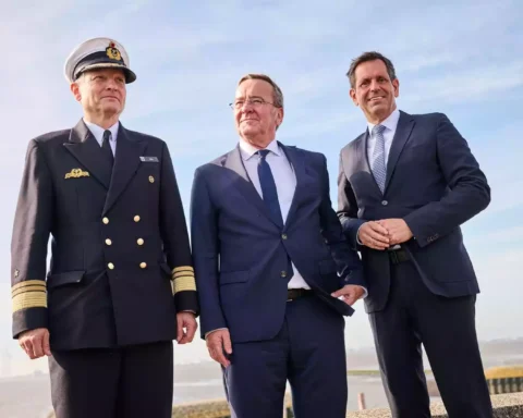 Drei Männer, darunter ein Marineoffizier und zwei Politiker in Anzügen, stehen am Wasser vor blauem Himmel.