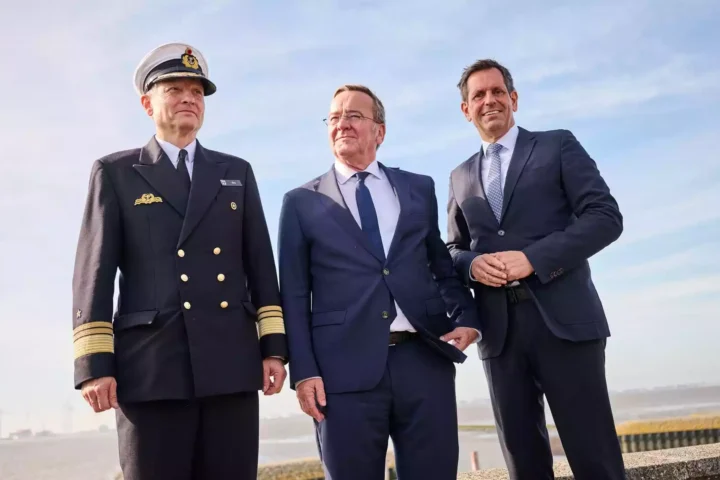 Drei Männer, darunter ein Marineoffizier und zwei Politiker in Anzügen, stehen am Wasser vor blauem Himmel.