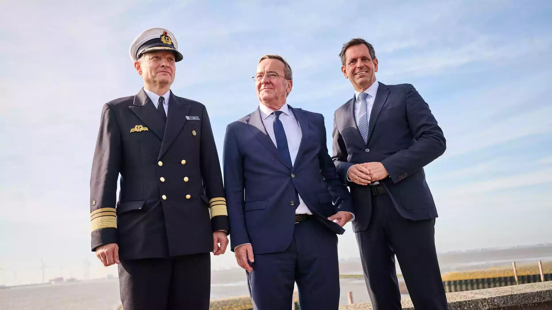 Bundeswehr plant zweiten Marinehafen in Emden 1 Drei Männer, darunter ein Marineoffizier und zwei Politiker in Anzügen, stehen am Wasser vor blauem Himmel.